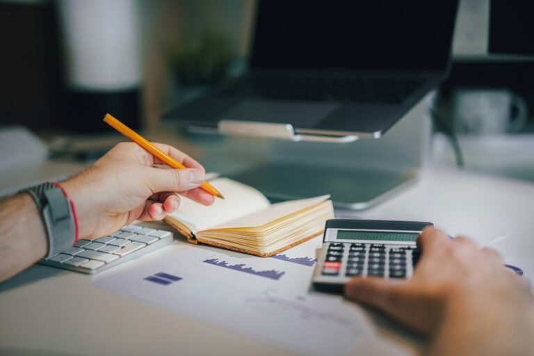 A person writing in a notebook next to a calculator and a laptop.