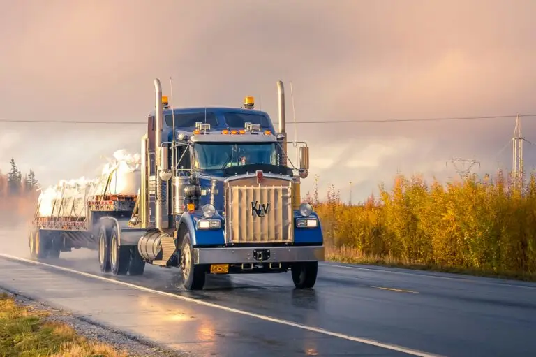 A blue semi truck driving on a wet road at sunrise.
