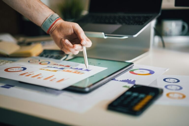 A hand using a stylus on a tablet showing charts, beside printed reports and a phone on a desk.