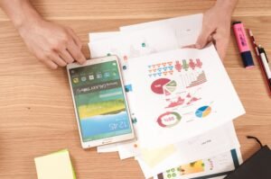 Hands on a wooden desk holding a phone beside printed pie and bar charts and sticky notes.